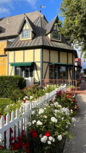 White picket fence with white and red flowers on each side with Danish style building in the background in Solvang,  CA